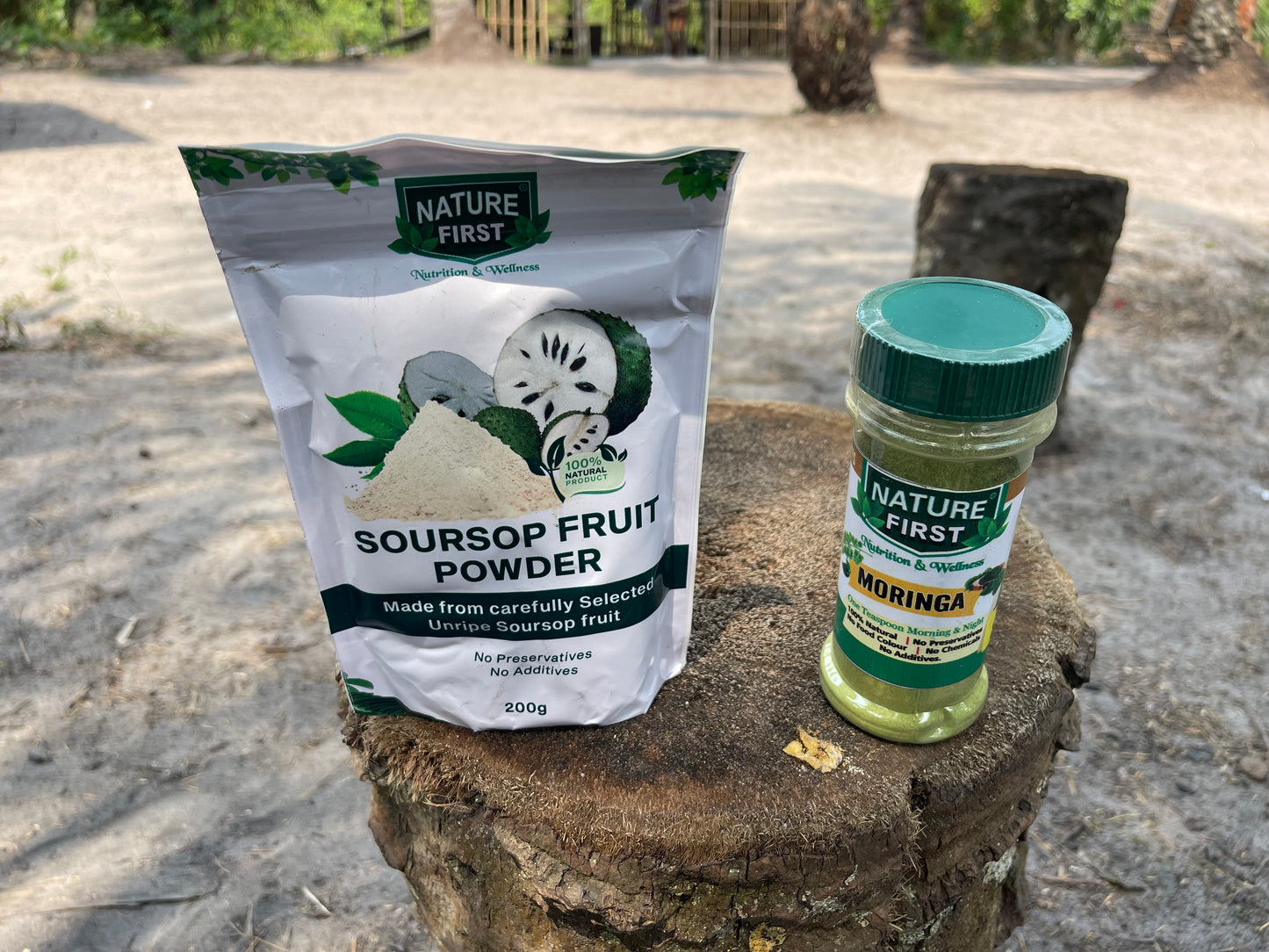 Nature First Soursop Fruit Powder package and Moringa bottle on a wooden stump outdoors.